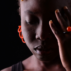 Frau mit leuchtend orangenem Ring und Ohrschmuck aus Karneol. Der Ring ist aus geschwärztem Silber mit einer Karneolkugel, der Ohrschmuck aus kleinen in Schlaufen gefädelten Karneolkügelchen gefertigt.