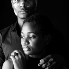 ringe_COUBO+carre Ein Mann steht hinter einer Frau. Beide tragen quadratische Ringe mit kristallisierten, quadratischen Onyx-Steinen besetzt.