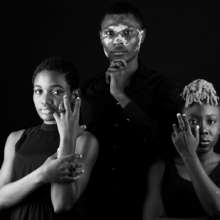 ringgruppe_carre+CUBO Ein Mann und zwei Frauen stehen in einer Gruppe und tragen eckige Ringe mit quadratischen Onyxsteinen besetzt.